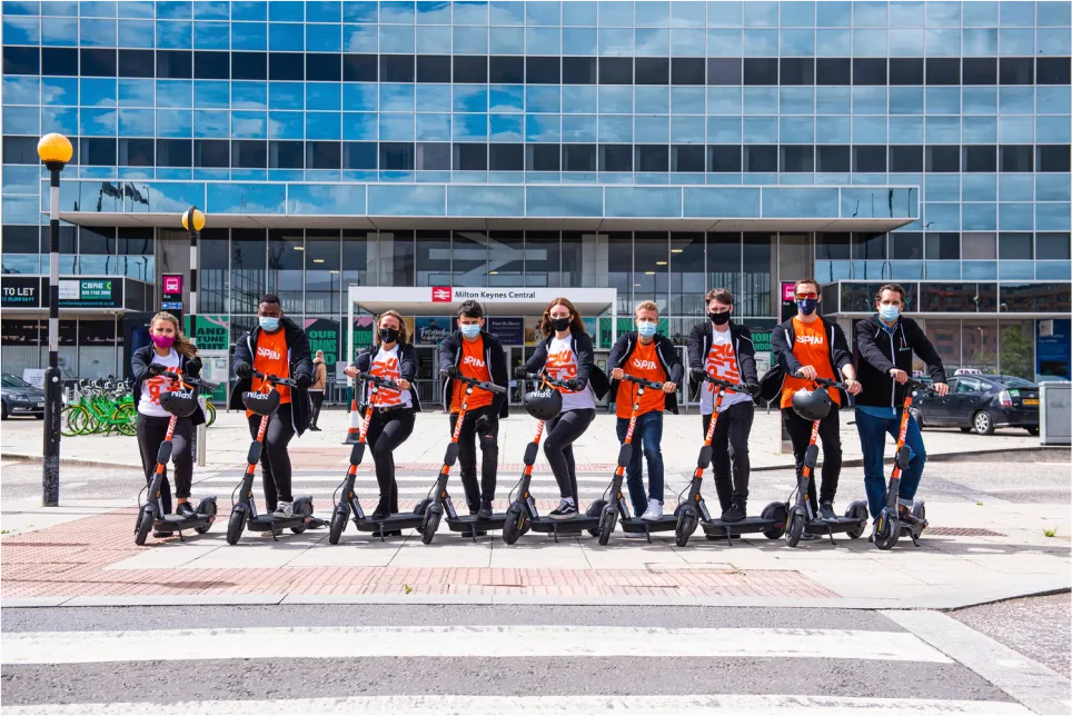 Many people posing with Spin Scooters outside a train station